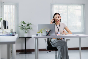 Young office worker sitting at her desk, wearing an ID card badge, engaging in a phone conversation while taking notes and working efficiently on her laptop in a bright, modern workspace