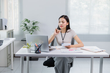 Young office worker sitting at desk and using laptop while holding calculator and reviewing documents, performing calculations and analyzing financial data for business strategy