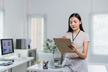 Young Asian businesswoman wearing an ID card and holding a clipboard, working diligently in a...