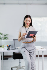 Young Asian businesswoman wearing lanyard with id card and using tablet, leaning on desk in modern office with laptop, smiling and looking at screen