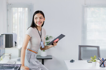 Young businesswoman is holding a tablet and smiling while sitting on her desk in a modern office,...