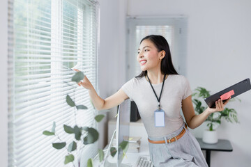 Young businesswoman is opening the blinds of her office window while holding a tablet, looking outside with a smile, enjoying the natural light and fresh air