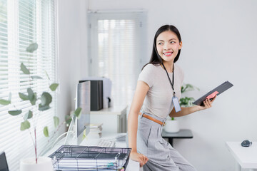 Young Asian businesswoman wearing gray trousers and beige t-shirt, holding tablet and smiling while leaning on desk in modern office with computer and blinds on window
