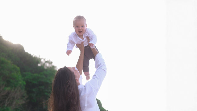 Portrait of a young woman walking on the beach with a baby in her arms. She throws the baby up in the air with the ocean in the background, the baby is surprised. Child first vacation with parents. 