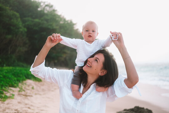 Portrait of mother and child who sits on her neck and laughs. Woman smiling and walking with child on the beach by the ocean. Vacation with little son. Mothers Day. - Powered by Adobe