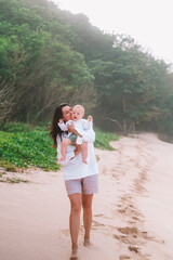 Portrait of a young woman walking on the beach with a baby in her arms. She hugs and kisses him on...
