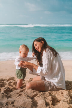 Portrait of mother and baby on vacation on the beach by the ocean. The little boy is capricious and crying, mother calms him down kisses and tickles him to cheer him up. Mothers Day. Travel with child