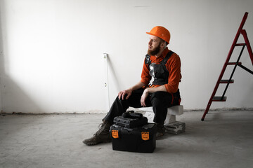 Builder or engineer in helmet and blue overalls sitting on construction site near toolbox looking...