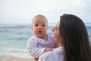 Close-up portrait of a baby with blue eyes being held by his mother against ocean. The mother is...