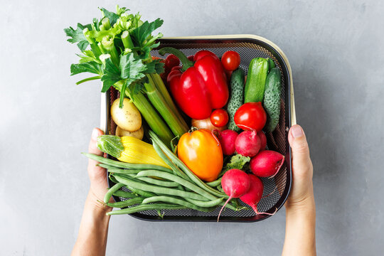 Woman's hands holding a basket of vegetables including tomatoes, radishes, and green beans, top view. The basket is filled with a variety of colorful vegetables