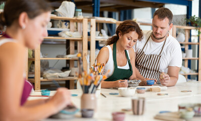 Student, in presence of guy master, paints bowl of gray clay, using brush to apply paint to surface...