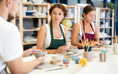 During working day, three pottery workshop workers sculpt from raw clay. They smooth surface of product with their fingers, form final product, and carry out order at desk.