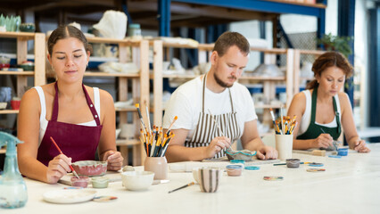 Three craftsperson are busy painting clay products. They are sitting near desktop, using brush and paints, painting product made of raw clay. Working together in pottery workshop.