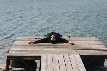Adult drunk man lying on wooden pier near water by sea, river outdoors with glass of alcohol...
