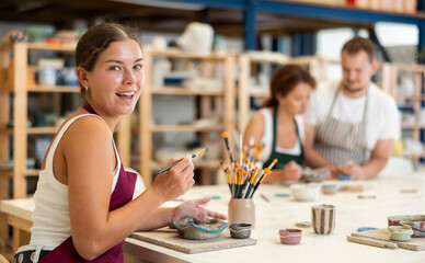 Concentrated girl in apron designing handmade clay plate with brush in taller room