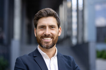 A smiling businessman in a suit, outdoors, with a building in the background.