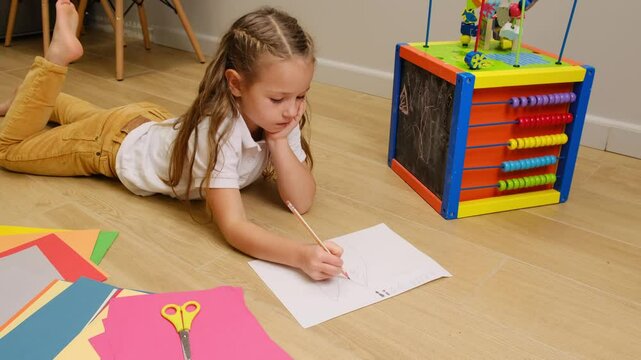 Cute little school child girl laying on floor making a painting sketching on paper using blue pen showing creative energy surrounded by toys at home