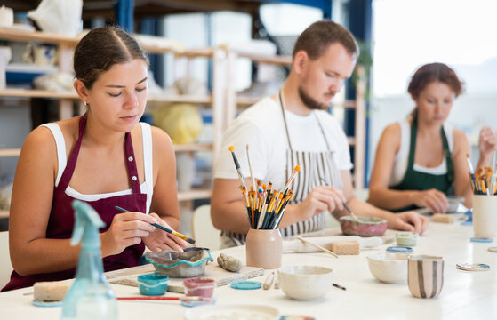 During working day, three employees of pottery workshop paint finished product from raw clay. They apply pattern to surface of plate with brush, dye decorative plate, carry out order at work table