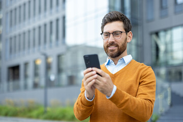 A businessman smiles while looking at his smartphone outside. He's wearing glasses and an orange...