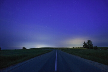 A deserted rural road stretches into the horizon under a starry night sky, illuminated by faint...
