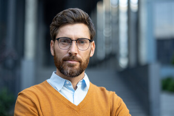 A portrait of a man with glasses and a beard, wearing a yellow sweater and a light blue shirt, taken outdoors.