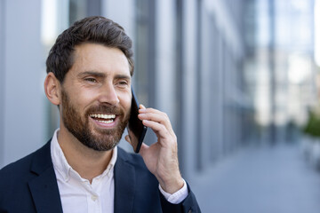 A smiling businessman in a suit, talking on a smartphone outdoors near a modern building.