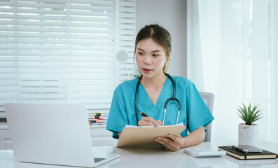 A female medical professional, who appears to be an East Asian female, in turquoise scrubs and a stethoscope, is actively working on a laptop while taking notes.