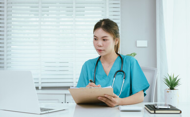 A female medical professional, who appears to be an East Asian female, in turquoise scrubs and a stethoscope, is actively working on a laptop while taking notes.