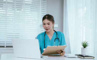 A female medical professional, who appears to be an East Asian female, in turquoise scrubs and a stethoscope, is actively working on a laptop while taking notes.