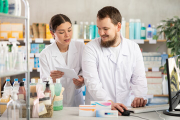 Obraz premium Man pharmacist at computer communicate with girl colleague. Employee discuss about treatment protocol, selects available drugs for treatment regimen