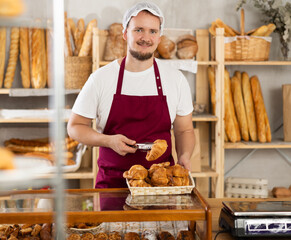 Young male salesperson lays out fresh croissants in showcase in bakery