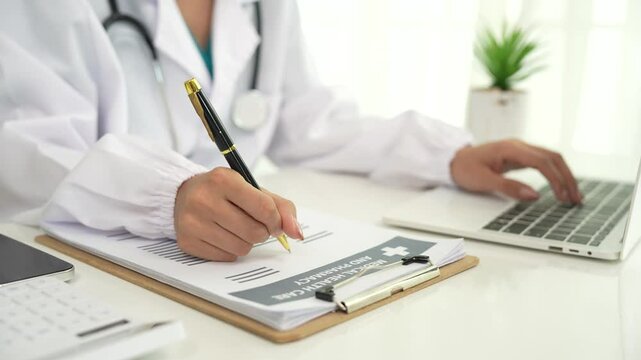 A professional in a white coat writes on a clipboard while using a laptop, suggesting healthcare administration, medical documentation, or online consultation.