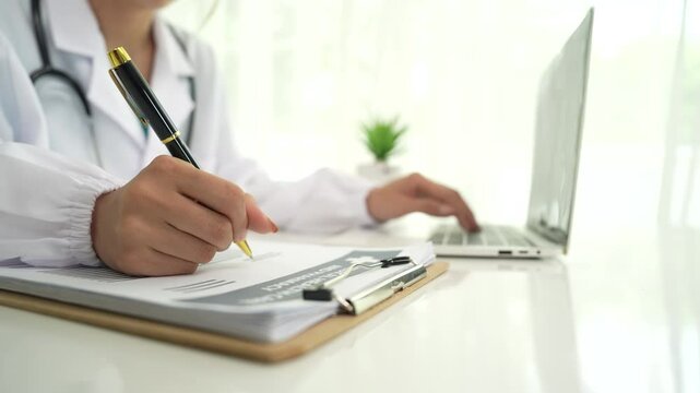 A professional in a white coat writes on a clipboard while using a laptop, suggesting healthcare administration, medical documentation, or online consultation.