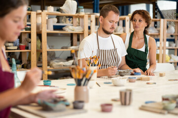 Student sits near desktop and paints clay product in presence of woman teacher. Lover of applied...