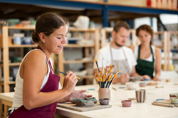 Female apprentice, is sitting near desktop and coloring clay product. In background, lovers of...