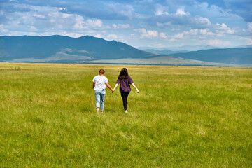 Two girls run through the green grass of the Barguzin Valley in the Republic of Buryatia. View from the back.