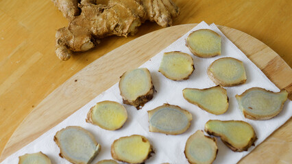 Thinly sliced pieces of blue ginger root are drying on a white paper towel placed on a wooden cutting board, with the rest of the ginger root visible in the background
