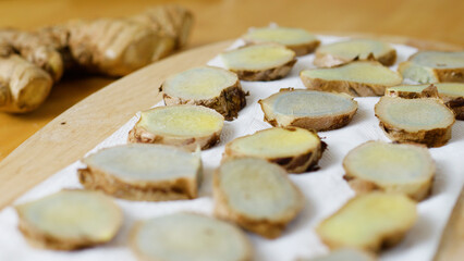 Freshly cut ginger root slices are drying on a white paper towel placed on a wooden cutting board, with the main root visible in the background, ready for use in culinary or medicinal applications