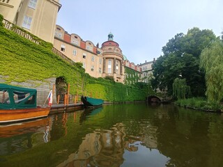 Boat is floating in a river next to a building. The boat is green and has a white flag on it....