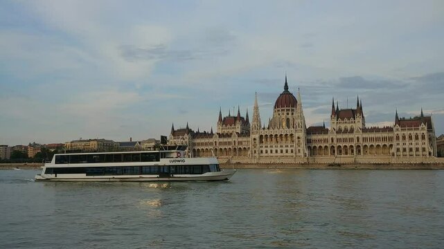 BUDAPESHT - JUL 21, 2019: Boat is in the water next to a large building Parliament. The building is a palace. The boat is blue. Budapest, Hungary