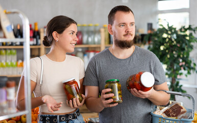 Couple looking at pickled vegetables in store. Market visitor choose consider jar with pickled...