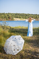 Woman with an umbrella in the summer heat. Woman in a blue dress standing by the lake. Blue water, beautiful nature and view