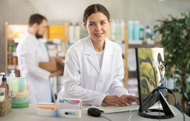 Young woman pharmacist with package of pills, she stands behind the counter near the computer....