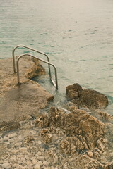 A rocky shoreline with a simple metal ladder leading into the calm, clear sea. Pebbles are visible...