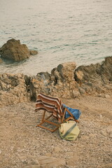 A wooden folding chair draped with a striped towel and a blue cloth stands on a rocky beach facing the calm sea.