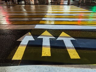 Colorful street markings reflect rain as pedestrians wait at a crosswalk in a bustling urban...