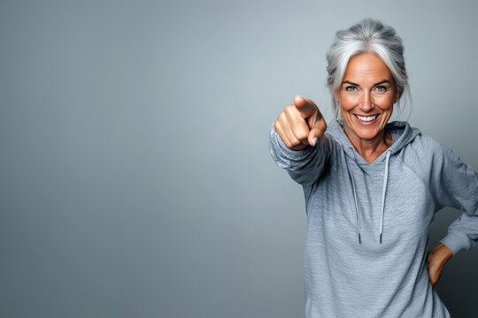 Smiling mature woman in sportswear pointing at camera over light grey background with copy space. Healthy lifestyle and fitness motivation concept - Powered by Adobe
