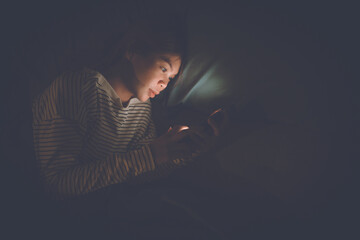 A young woman lies awake at night in bed, scrolling on her glowing phone, battling insomnia,...