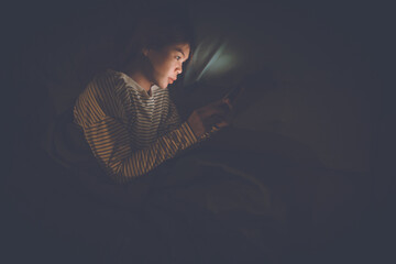 A young woman lies awake at night in bed, scrolling on her glowing phone, battling insomnia,...