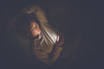 A young woman lies awake at night in bed, scrolling on her glowing phone, battling insomnia,...